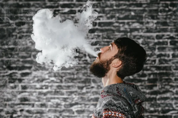 Un joven con barba vapeando un cigarrillo electrónico. Vaporizador de humo Vaper hipster . — Foto de Stock