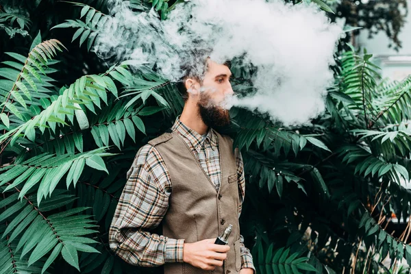 Joven con barba vapeando un cigarrillo electrónico al aire libre. vaporizador de humo hipster . — Foto de Stock