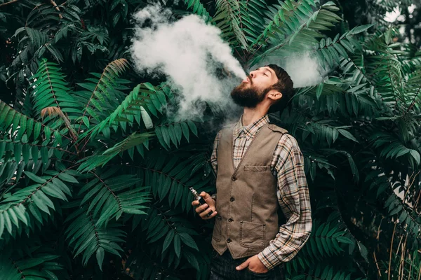 Joven con barba vapeando un cigarrillo electrónico al aire libre. vaporizador de humo hipster . — Foto de Stock