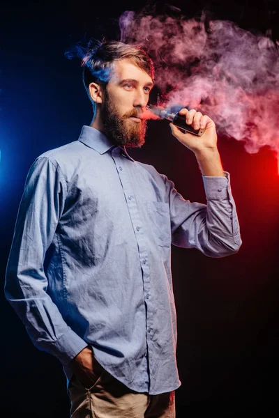 Un joven con barba vapeando y libera una nube de vapor. vaporizador de humo hipster vaper . — Foto de Stock