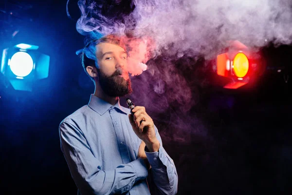 Un joven con barba vapeando y libera una nube de vapor. vaporizador de humo hipster vaper . — Foto de Stock