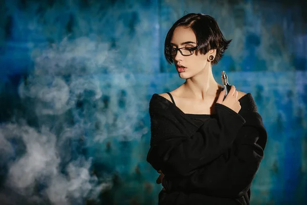 Девушка в очках испаряется и выпускает облако пара. Модель в черном испарителе дыма на бирюзовом фоне — стоковое фото