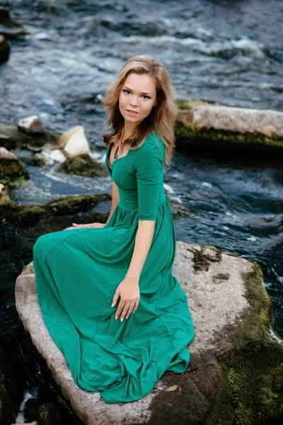 Piękna dziewczyna pozuje na skały w pobliżu wody. Model w zieloną sukienkę w przyrodzie. — Zdjęcie stockowe
