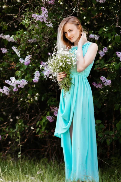 Blond tjej med bukett av vilda blommor utomhus en modell i en turkos klänning mot bakgrund av en lila buske. — Stockfoto