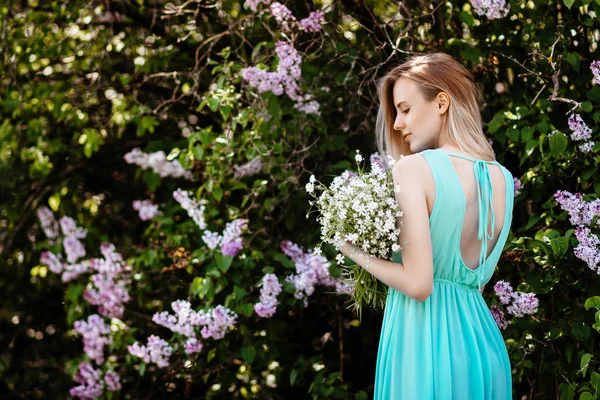 Blondýnka s kytice divokých květin venku A modelu v tyrkysových šatech pozadí lila Bushe. — Stock fotografie