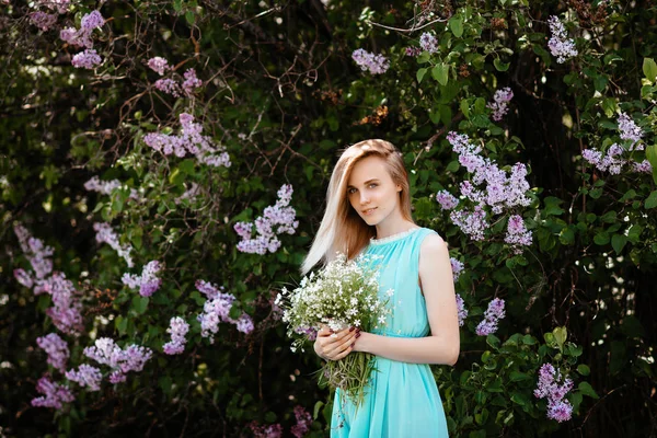Chica rubia con ramo de flores silvestres al aire libre Un modelo en un vestido de color turquesa sobre un fondo de un arbusto lila . — Foto de Stock