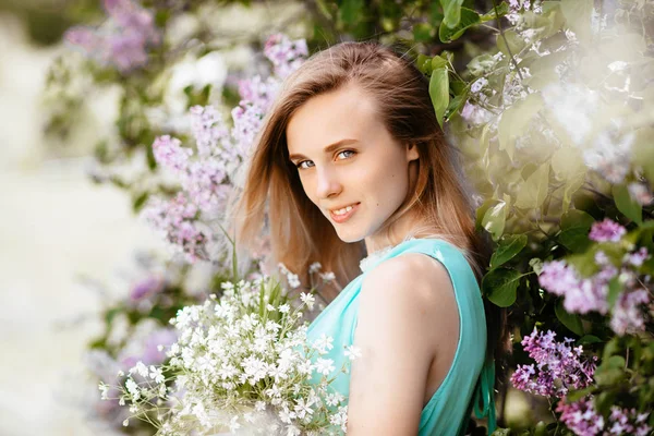 ライラック ブッシュの背景に青緑色のドレスでアウトドア モデルを野生の花の花束とブロンドの女の子. — ストック写真