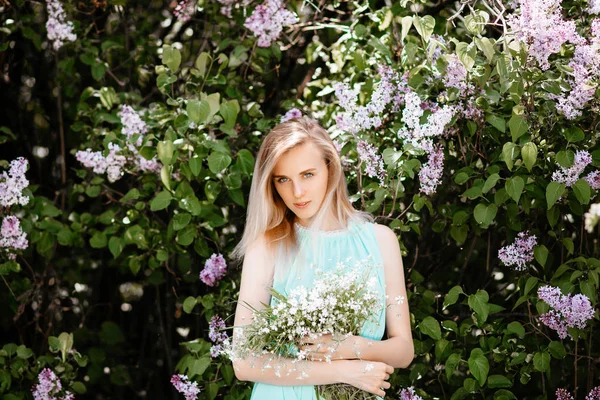 Блондинка з букетом з диких квітів на відкритому повітрі Модель в бірюзовій сукні на фоні бузкового куща . — стокове фото