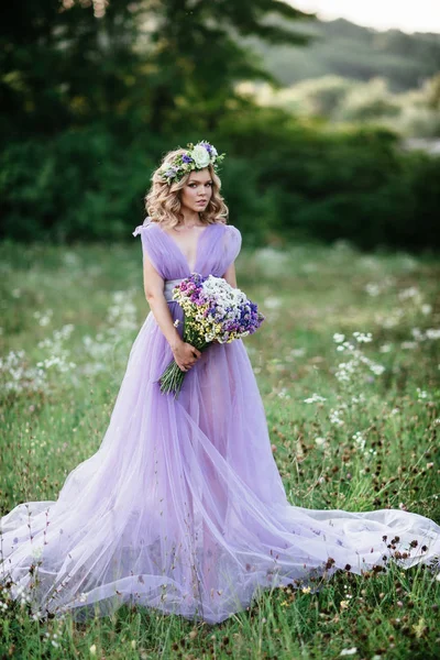 Schoonheid vrouw portret met krans van bloemen op hoofd. bruid in paarse jurk met boeket van wilde bloemen. Buiten. Soft Focus Sea... — Stockfoto