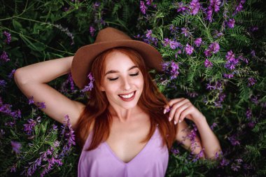 Şapkalı kızıl saçlı kız elinde mor çiçeklerle çimlerin üzerinde yatıyor. Genç kadın doğada gülümsüyor..