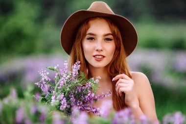 Şapkalı kızıl saçlı kız elinde mor çiçeklerle çimlerin üzerinde yatıyor. Genç bir kadın doğada gülümser. Bayan lavanta tarlasında yürüyor..