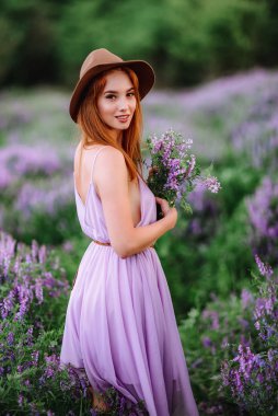Şapkalı kızıl saçlı kız elinde mor çiçeklerle çimlerin üzerinde yatıyor. Genç bir kadın doğada gülümser. Bayan lavanta tarlasında yürüyor..