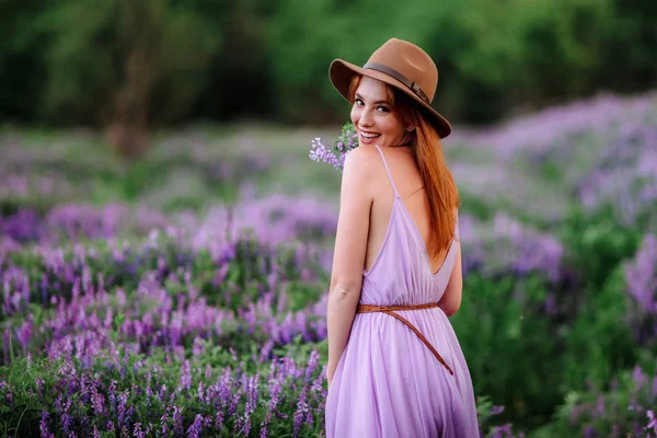 帽子の赤い髪の女の子は紫色の花を持つ草の中にあります。若い女性は自然に笑顔。女性はラベンダー畑を歩き. — ストック写真