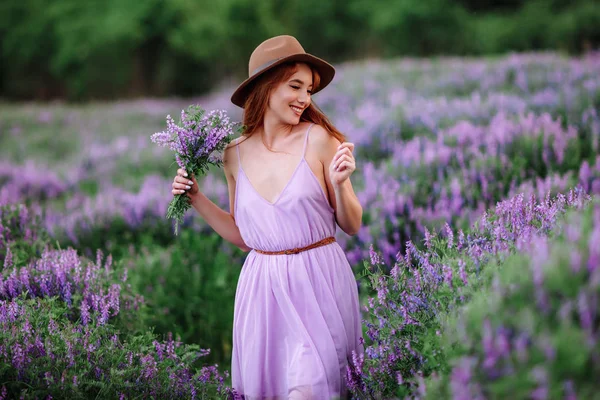 帽子の赤い髪の女の子は紫色の花を持つ草の中にあります。若い女性は自然に笑顔。女性はラベンダー畑を歩き. — ストック写真