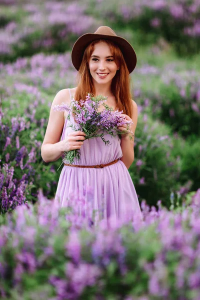Red-haired girl in a hat lies in the grass with purple flowers. Young woman smile in nature. lady walks on a lavender field. — Stock Photo, Image
