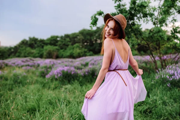 Rudowłosa dziewczyna w kapeluszu leży w trawie z fioletowymi kwiatami. Młoda kobieta uśmiecha się w naturze. pani spacery na polu lawendy. — Zdjęcie stockowe