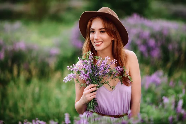 帽子の赤い髪の女の子は紫色の花を持つ草の中にあります。若い女性は自然に笑顔。女性はラベンダー畑を歩き. — ストック写真