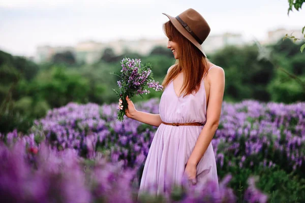 Červenovlasá dívka v klobouku leží v trávě s fialovými květy. Mladá žena se v přírodě usmívá. dáma kráčí po levandulovém poli. — Stock fotografie