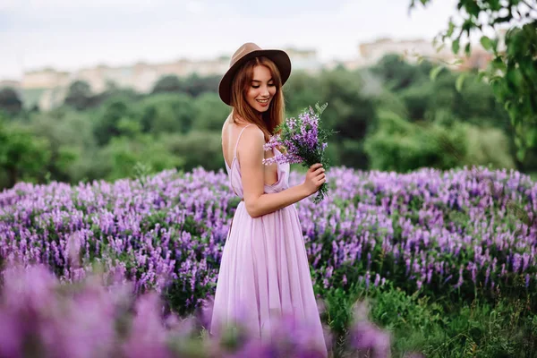 帽子の赤い髪の女の子は紫色の花を持つ草の中にあります。若い女性は自然に笑顔。女性はラベンダー畑を歩き. — ストック写真