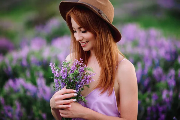 帽子の赤い髪の女の子は紫色の花を持つ草の中にあります。若い女性は自然に笑顔。女性はラベンダー畑を歩き. — ストック写真