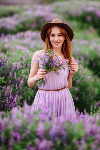 帽子の赤い髪の女の子は紫色の花を持つ草の中にあります。若い女性は自然に笑顔。女性はラベンダー畑を歩き. — ストック写真