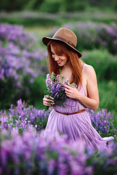 帽子の赤い髪の女の子は紫色の花を持つ草の中にあります。若い女性は自然に笑顔。女性はラベンダー畑を歩き. — ストック写真