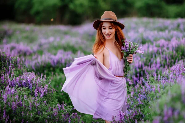 Červenovlasá dívka v klobouku leží v trávě s fialovými květy. Mladá žena se v přírodě usmívá. dáma kráčí po levandulovém poli. — Stock fotografie