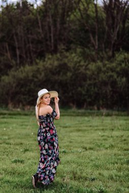 Yeşil bir çayırda, elbise ve şapkalı güzel bir kız tek başına yürüyor. Genç ve güzel sarışın kadın özgürlüğün ve rahatlığın tadını çıkarıyor.
