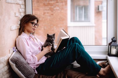Gözlüklü kız pencerenin yanında bir kitap okuyor. Genç bir kadın, kedisiyle balkonda dinleniyor.