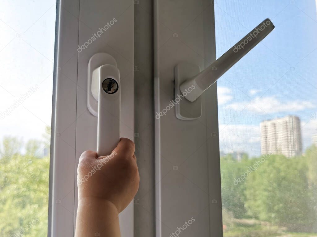 A children's hand is trying to open a window with a lock on the handle in an apartment on a high floor. A security lock for windows ensures the safety of your children and prevents accidents.