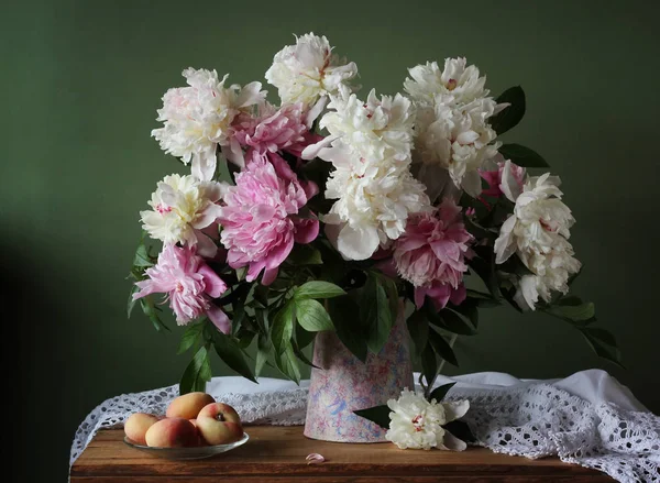 Boeket van pioenrozen in een kruik en perziken op tafel. — Stockfoto