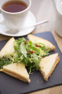 Beyaz bir tabakta servis edilen çeşitli kulüp sandviçleri bir fincan sıcak çay ve demlikte. Lezzetli