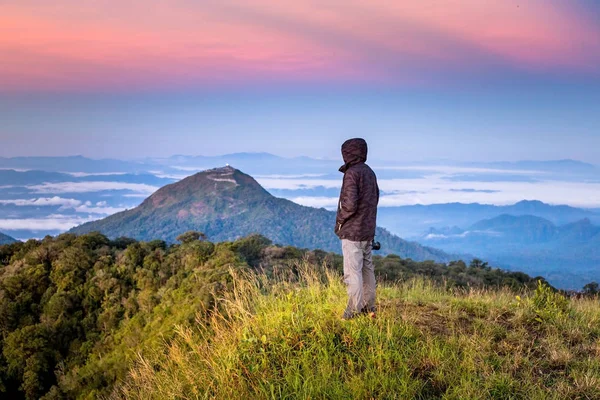 Ο άνθρωπος στην κουκούλα στέκεται στην κορυφή του βουνού με την όμορφη ανατολή του ήλιου — Φωτογραφία Αρχείου