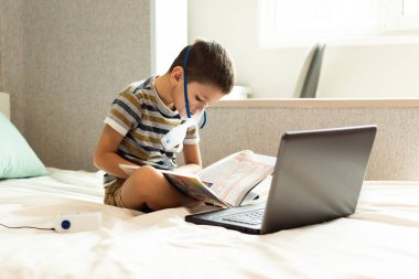 Evde kalıp koronavirüs salgınını önleyin. Bir çocuk karantina sırasında oksijen maskesi, dizüstü bilgisayar ve kitapla evde ders alır. Önleme salgını. COVID-19.