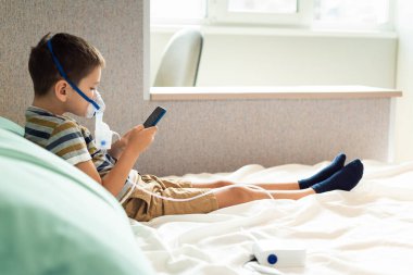 Evde kalıp koronavirüs salgınını önleyin. Karantina sırasında cep telefonuyla oynayan oksijen maskeli bir çocuk. Önleme salgını. COVID-19.