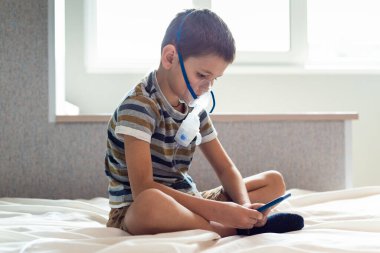 Evde kalıp koronavirüs salgınını önleyin. Karantina sırasında cep telefonuyla oynayan oksijen maskeli bir çocuk. Önleme salgını. COVID-19.