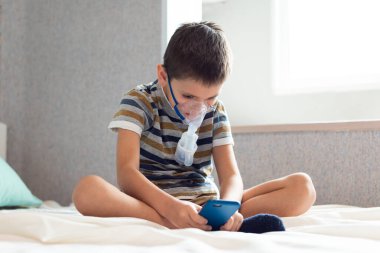 Evde kalıp koronavirüs salgınını önleyin. Karantina sırasında cep telefonuyla oynayan oksijen maskeli bir çocuk. Önleme salgını. COVID-19.