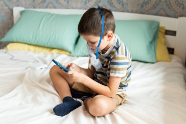 Evde kalıp koronavirüs salgınını önleyin. Karantina sırasında cep telefonuyla oynayan oksijen maskeli bir çocuk. Önleme salgını. COVID-19.