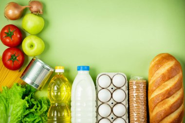 Temel taze ürünler yeşil arka planda yatar. Yumurta, yeşillik, spagetti, domates, ekmek, süt, kurabiye, konserve yiyecek. Yemek teslimatı covid-19 salgını, bağış. Üst Manzara. Boşluğu kopyala.