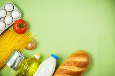 Temel taze ürünler yeşil arka planda yatar. Yumurta, yeşillik, spagetti, domates, ekmek, süt, kurabiye, konserve yiyecek. Yemek teslimatı covid-19 salgını, bağış. Üst Manzara. Boşluğu kopyala.