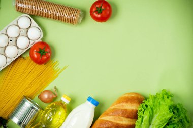 Temel taze ürünler yeşil arka planda yatar. Yumurta, yeşillik, spagetti, domates, ekmek, süt, kurabiye, konserve yiyecek. Yemek teslimatı covid-19 salgını, bağış. Üst Manzara. Boşluğu kopyala.