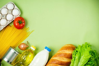 Temel taze ürünler yeşil arka planda yatar. Yumurta, yeşillik, spagetti, domates, ekmek, süt, kurabiye, konserve yiyecek. Yemek teslimatı covid-19 salgını, bağış. Üst Manzara. Boşluğu kopyala.