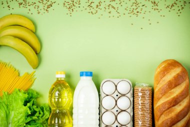 Temel taze ürünler yeşil arka planda yatar. Yumurta, yeşillik, spagetti, ekmek, süt, tuvalet kağıdı, kurabiye, ayçiçeği yağı. Yemek teslimatı covid-19 salgını, bağış. Üst Manzara. Boşluğu kopyala.