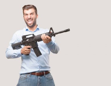 genç yakışıklı adam kızgın ifade bir mavi t gömlek giymiş bir tüfek tutarak. Tek renkli arka planı izole kişi.