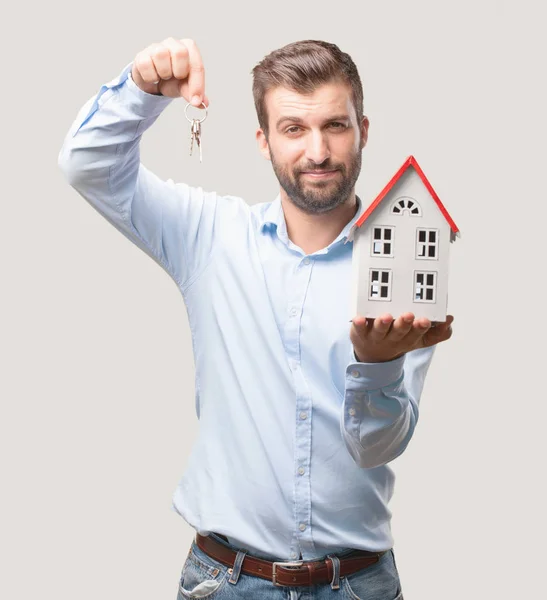 年轻英俊的男子拿着一个房子模型和钥匙穿着蓝色 独立于单色背景的人 — 图库照片