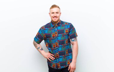 Havalı tişörtlü genç sarışın adam mutlu bir şekilde gülümsüyor, eli kalçasında ve kendinden emin, pozitif, gururlu ve beyaz arka plana karşı dost canlısı bir tutum sergiliyor.