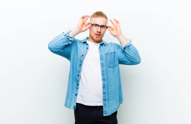 Kot pantolon giyen genç sarışın adam şok olmuş, şaşırmış ve şaşırmış hissediyor. Beyaz arka planda hayrete düşmüş, inançsız bir bakış atıyor.