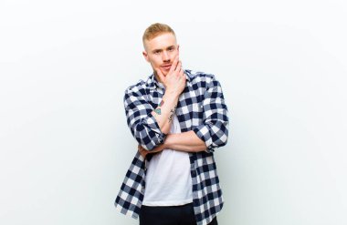 Kare gömlekli genç sarışın adam ciddi, düşünceli ve güvensiz görünüyor, bir kolu çapraz ve çenesi üzerinde, seçenekleri tartıyor.