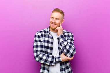 Kare gömlekli genç sarışın adam mutlu bir şekilde gülümsüyor ve hayal kuruyor ya da şüphe ediyor.