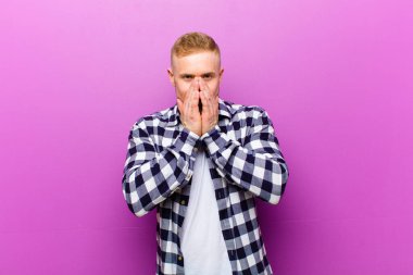 Kare gömlekli genç sarışın adam mutlu, neşeli, şanslı ve şaşkın görünüyor iki eliyle ağzını kapatıyor.